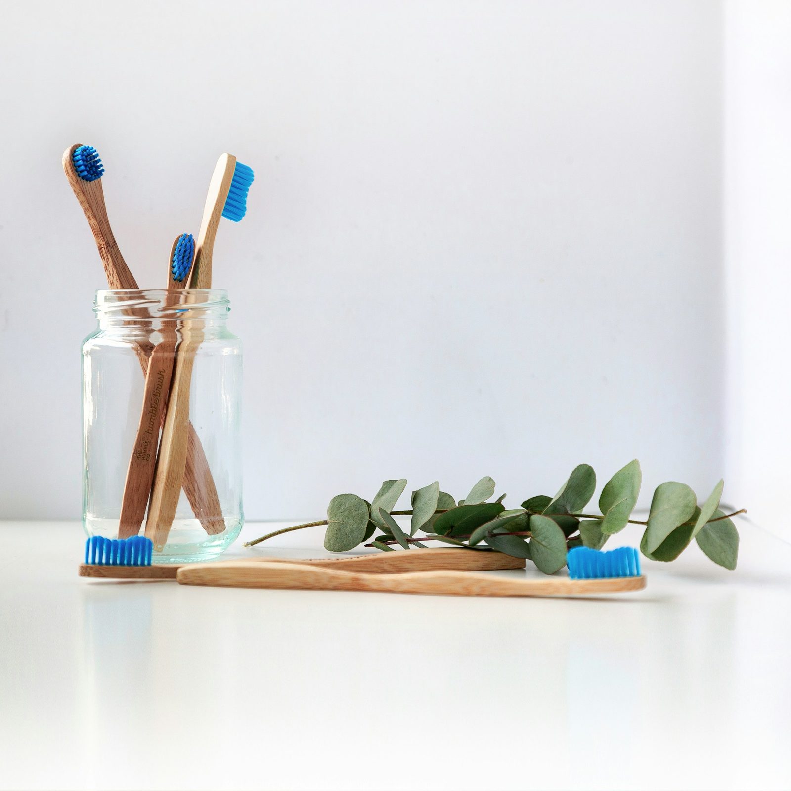 Bamboo toothbrushes in a glass jar beside fresh green leaves on a bright white counter